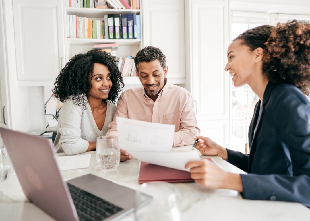 “Photograph of a smiling couple discussing home-buying paperwork with an agent” - Source: Canva