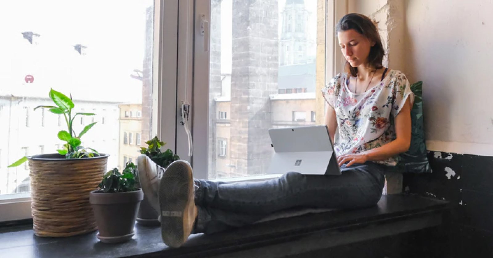  Woman Seated Beside Window on Her Laptop with Three Plants at Her Feet with Cityscape in Background