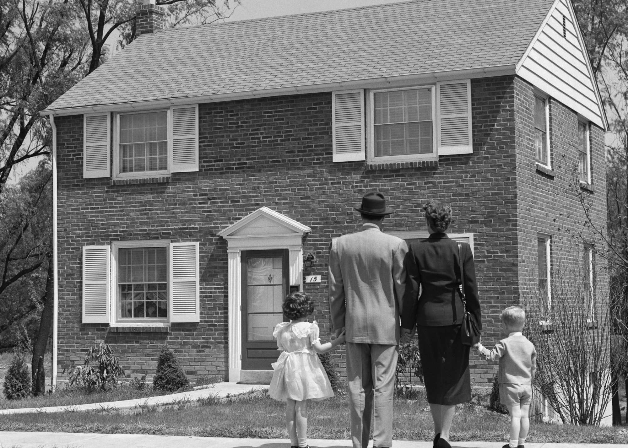 Black-and-white photo of a family of 4 standing in front of a two-story brick home - Armstrong Roberts/ClassicStock // Getty Images