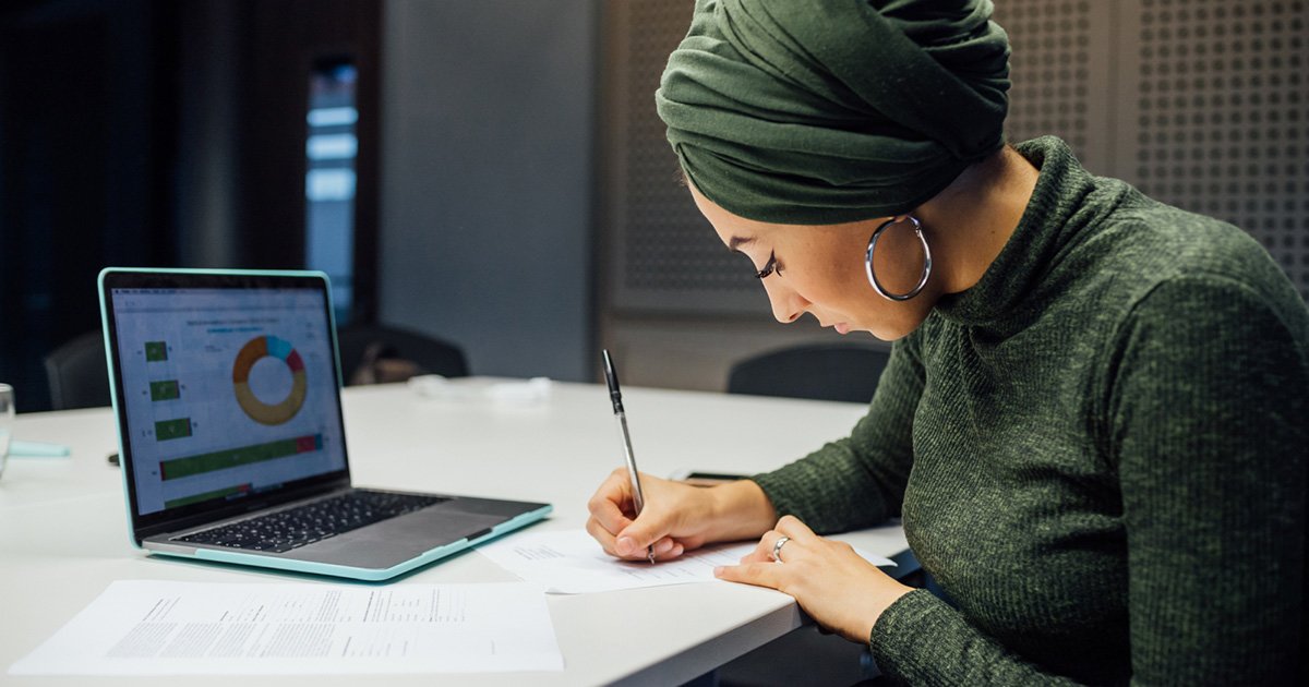 Woman in Green Top and Headwrap Writing on White Table in Front of an Open Laptop