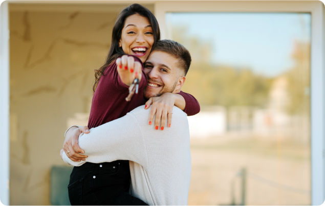 Happy couple celebrating outside their new home, with the woman holding a key and being lifted in the man&#39;s arms.