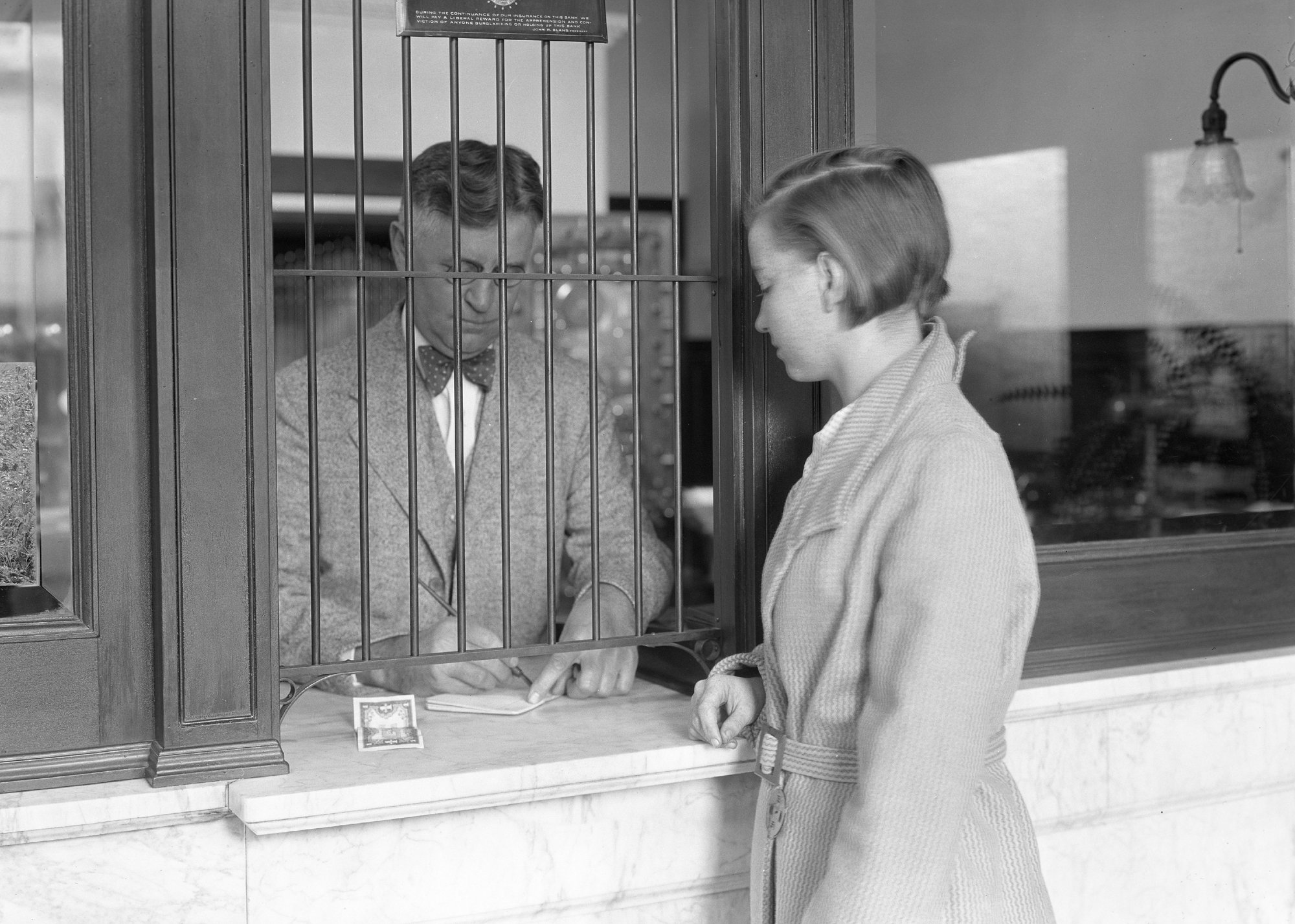 Black-and-white photo of a woman conducting a transaction at a bank teller’s counter - PhotoQuest // Getty Images