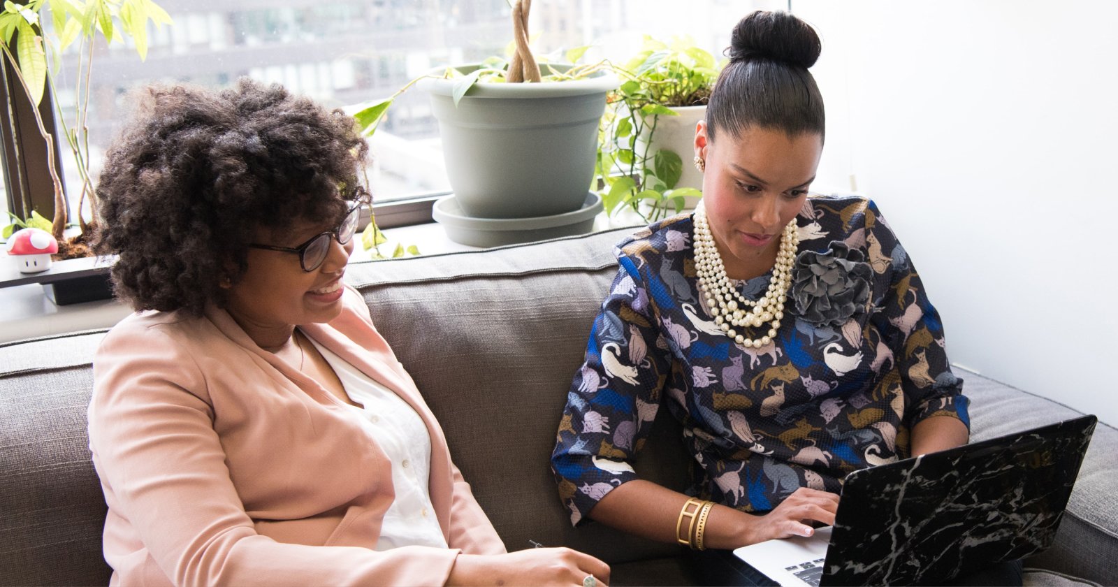 Two women sitting on a couch in a sunny room reviewing documents on a laptop