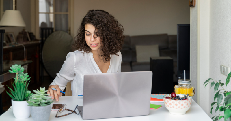 Person Sitting at Desk with Laptop, Pen In Hand, Viewing Cellular Phone