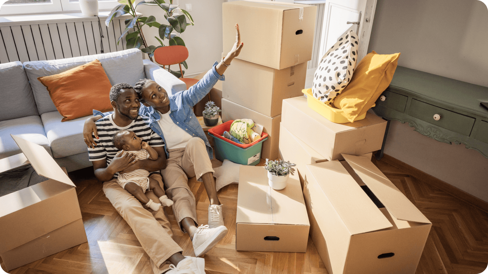 Happy family sitting on the floor of their new home, surrounded by moving boxes, with a baby in their arms and the mother waving cheerfully.