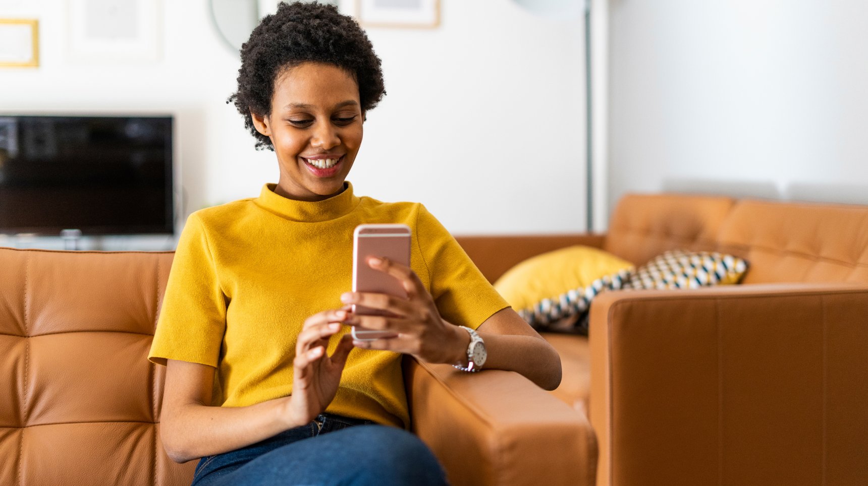 Smiling Woman in Mustard Top on a Cell Phone Sitting on a Caramel Leather Couch