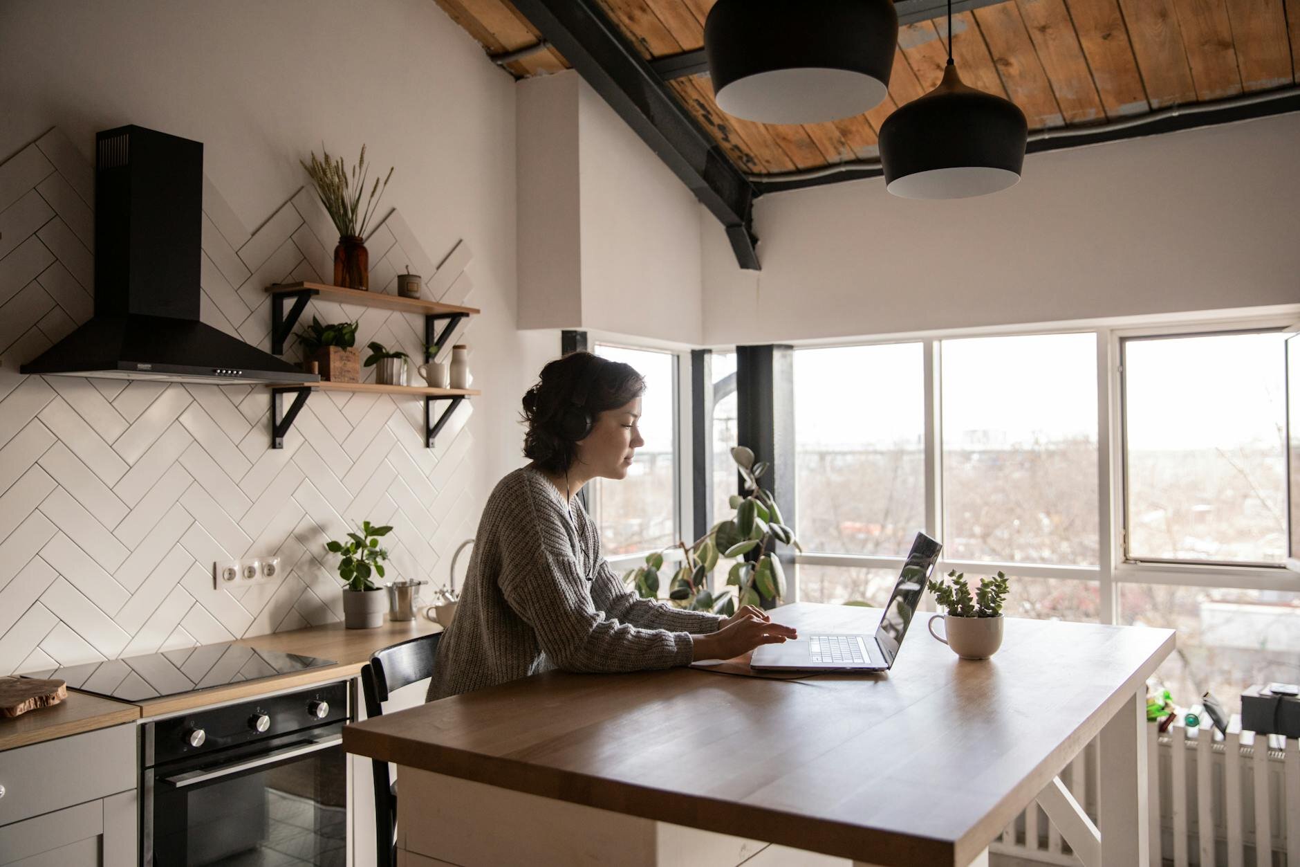 Person sitting at a kitchen island working on a laptop with a modern open-ceiling kitchen and large windows in the background.