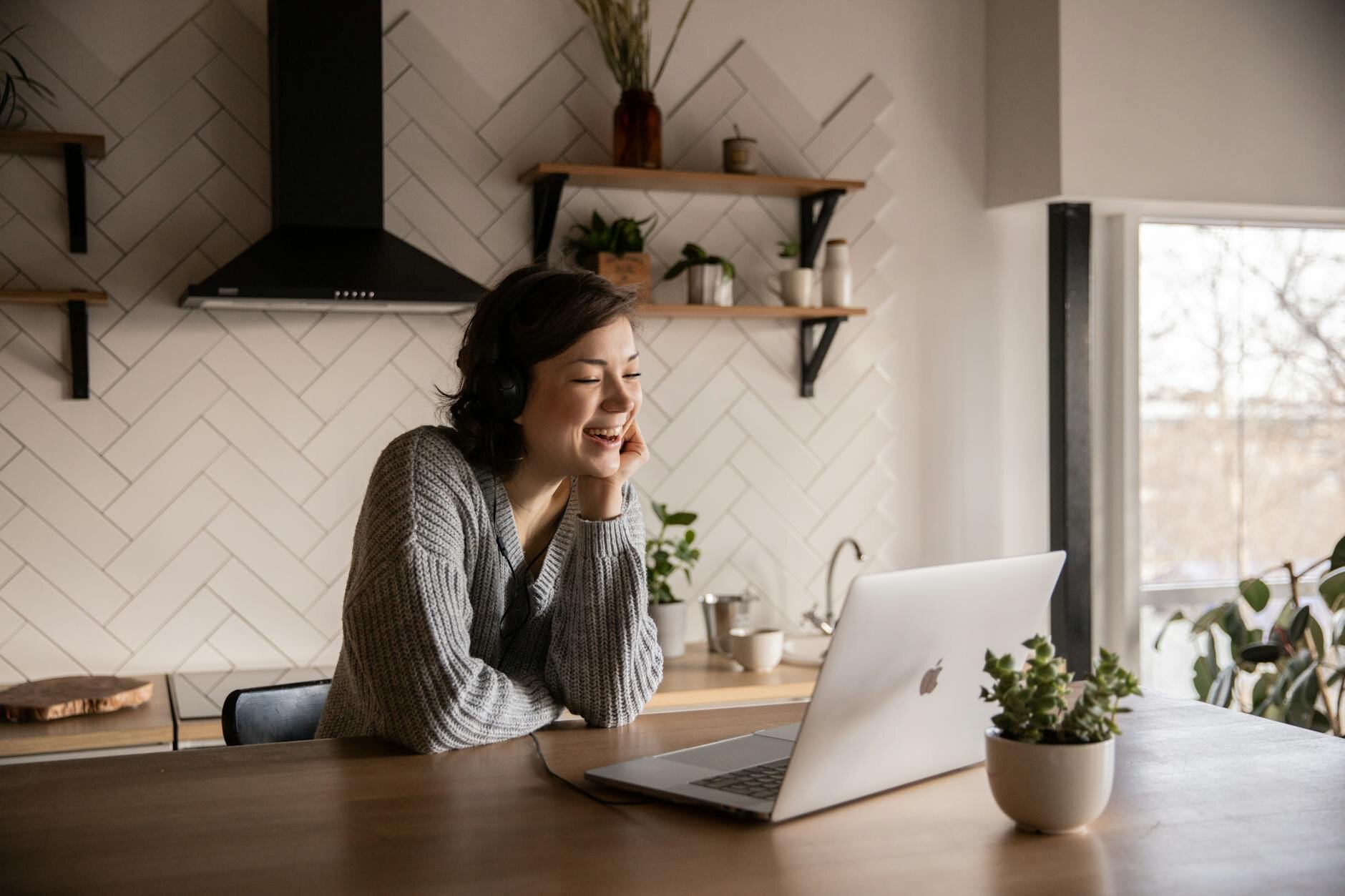Woman wearing headphones smiling at a laptop in a cozy kitchen.