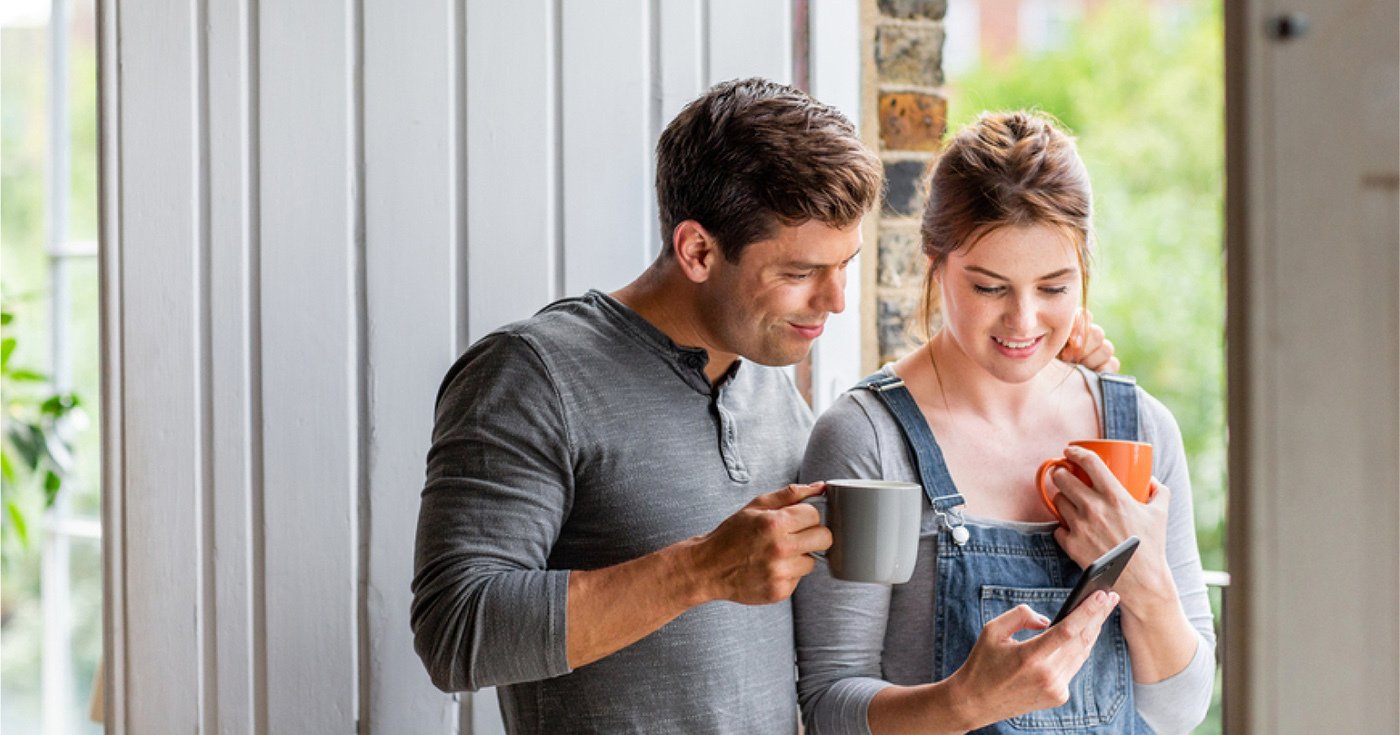 Embracing Couple Holding Mugs Showing Each Other Something on a Cell Phone