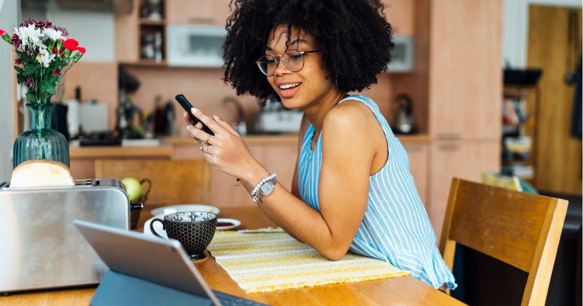 Person Sitting at Wooden Kitchen Table Looking at their Laptop with Cell Phone in Hand