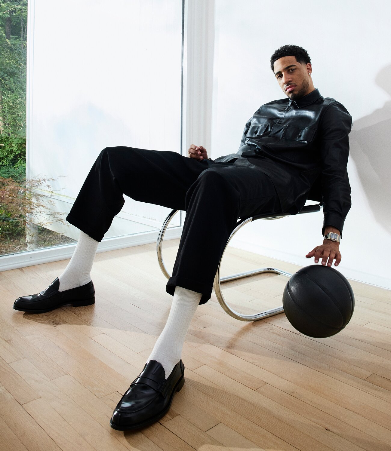 Tyrese Haliburton sitting in a chair, bouncing a black basket ball, wearing a black leather jacket, black pants, high white socks, black loafers and the Heritage 1917 watch with stainless steel bracelet and grey dial.