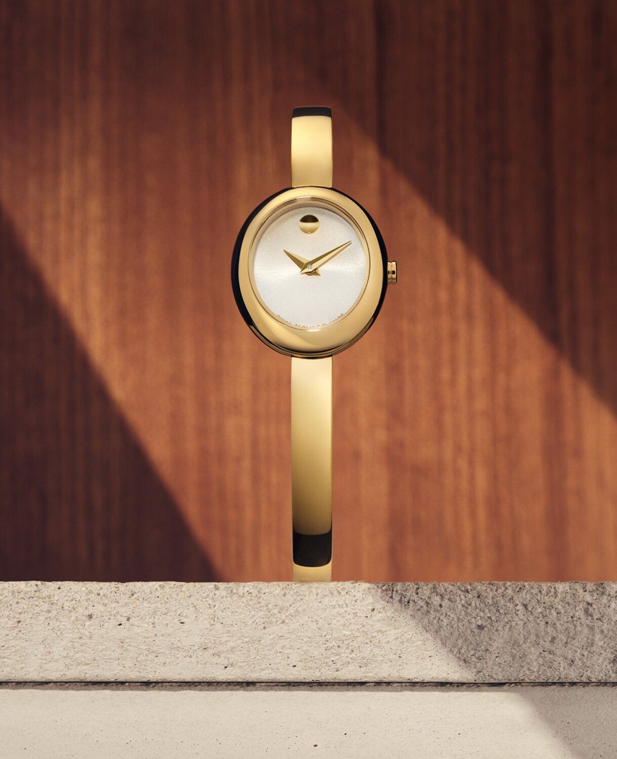 Museum Bangle watch with gold tone bracelet and white oval dial sitting on stone in front of a wooden panel.