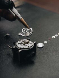 Tyrese Haliburton sitting in a chair, bouncing a black basket ball, wearing a black leather jacket, black pants, high white socks, black loafers and the Heritage 1917 watch with stainless steel bracelet and grey dial.