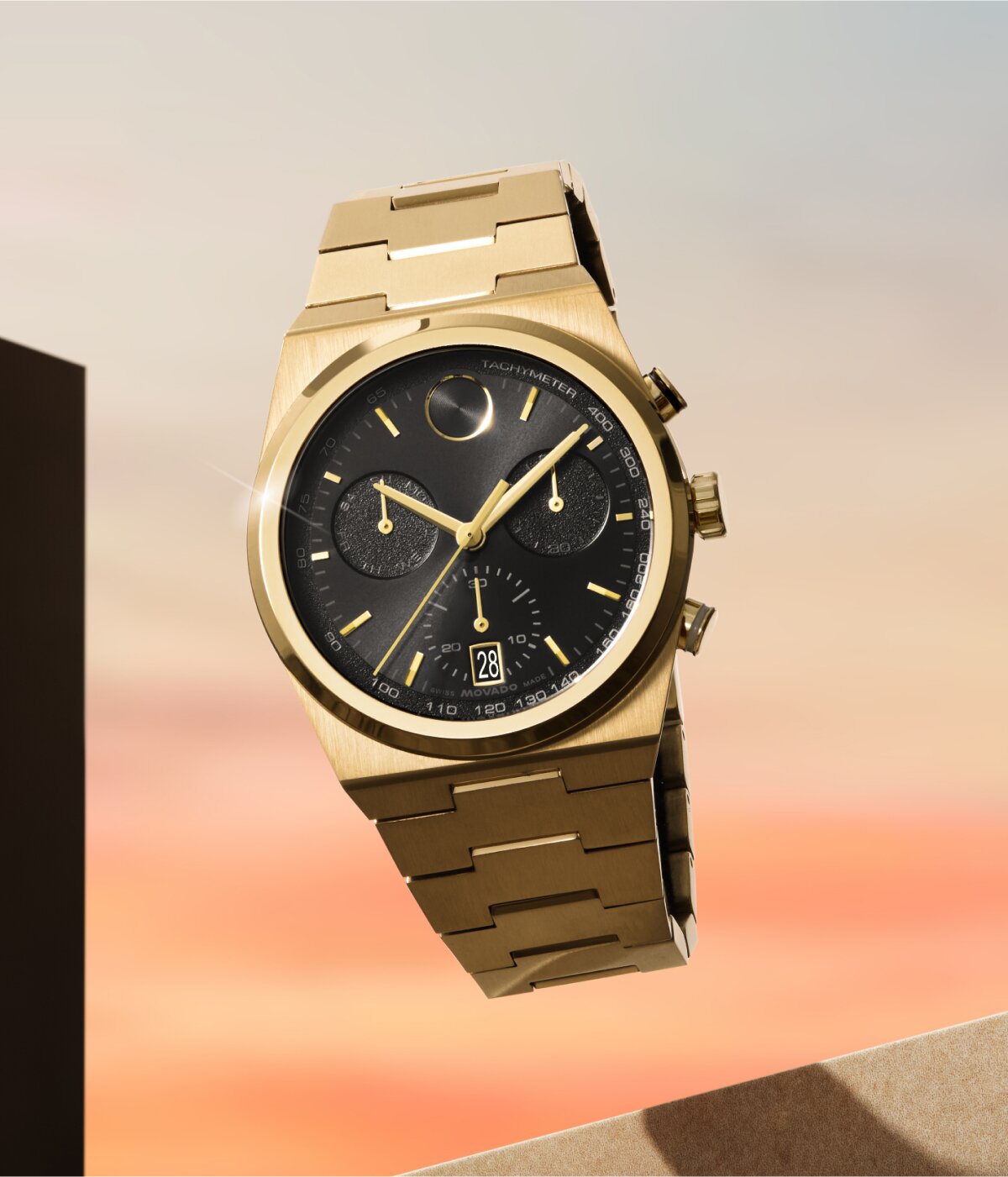 Movado BOLD Quest Chronograph with gold bracelet and black dial floating in front of a sunset.