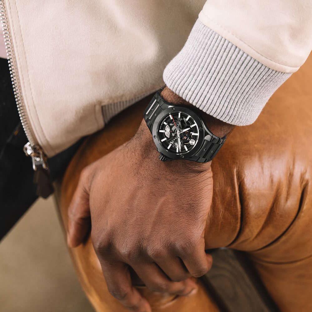 Man leaning on a brown leather couch wearing a jacket and a Black plated stainless steel Alta SE watch with skeleton dial.