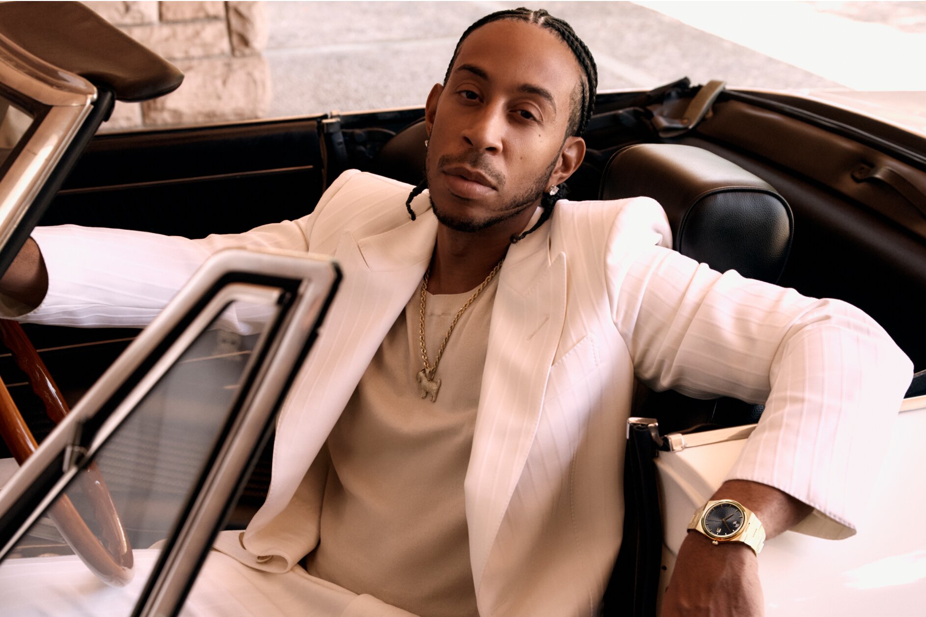 Ludacris sitting in a car, wearing a white suite and the BOLD Quest watch with gold tone bracelet and black dial.