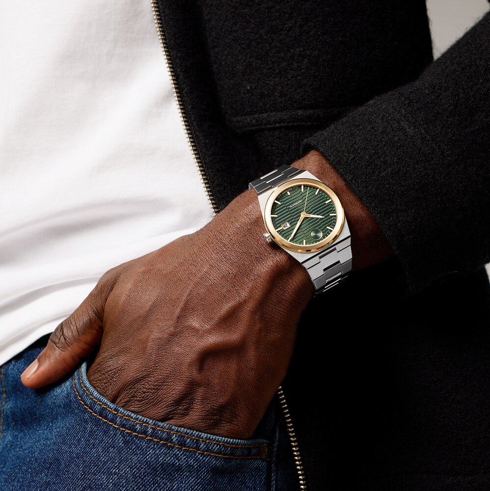 Man in a grey sweater with his hand on his shoulder wearing the Faceto watch with gold tone bracelet and black dial with diamonds.