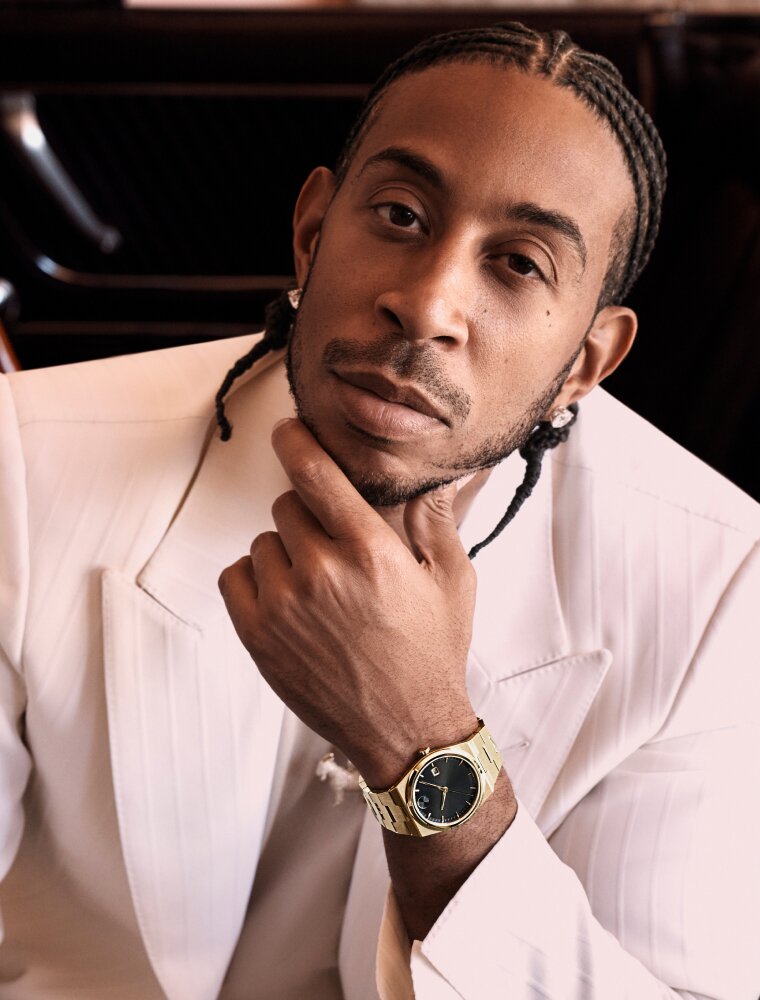 Ludacris wearing a white suit, sitting in a car, wearing the BOLD Quest watch with gold tone bracelet and black dial.