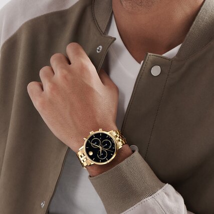 Man in a green jacket wearing the Museum Classic watch with gold tone bracelet and black chronograph dial.