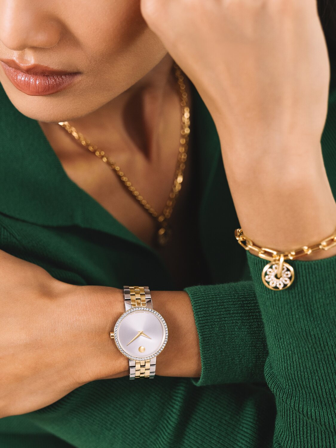 Woman in a green sweater wearing a two tone Museum Classic bracelet watch featuring lab grown diamonds and a gold tone fleur necklace and bracelet that features lab grown diamonds.