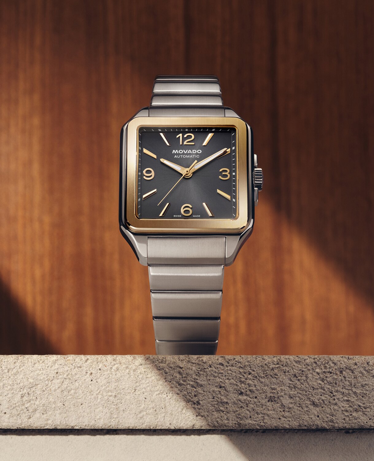 Heritage 1917 watch with stainless steel bracelet and grey dial sitting on stone in front of a wooden wall.