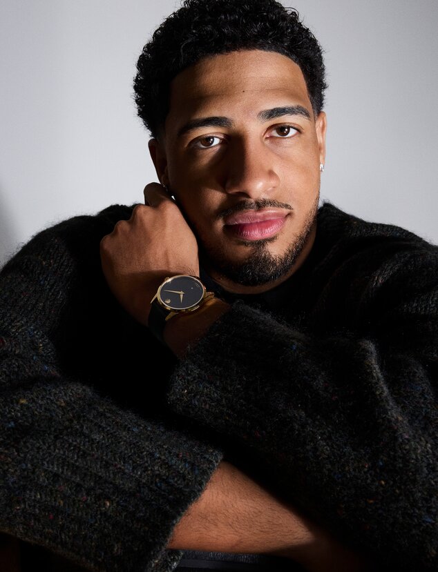Tyrese Haliburton sitting in front of a grey background wearing a black sweater and the Museum Classic automatic watch with black strap and dial and gold accents.