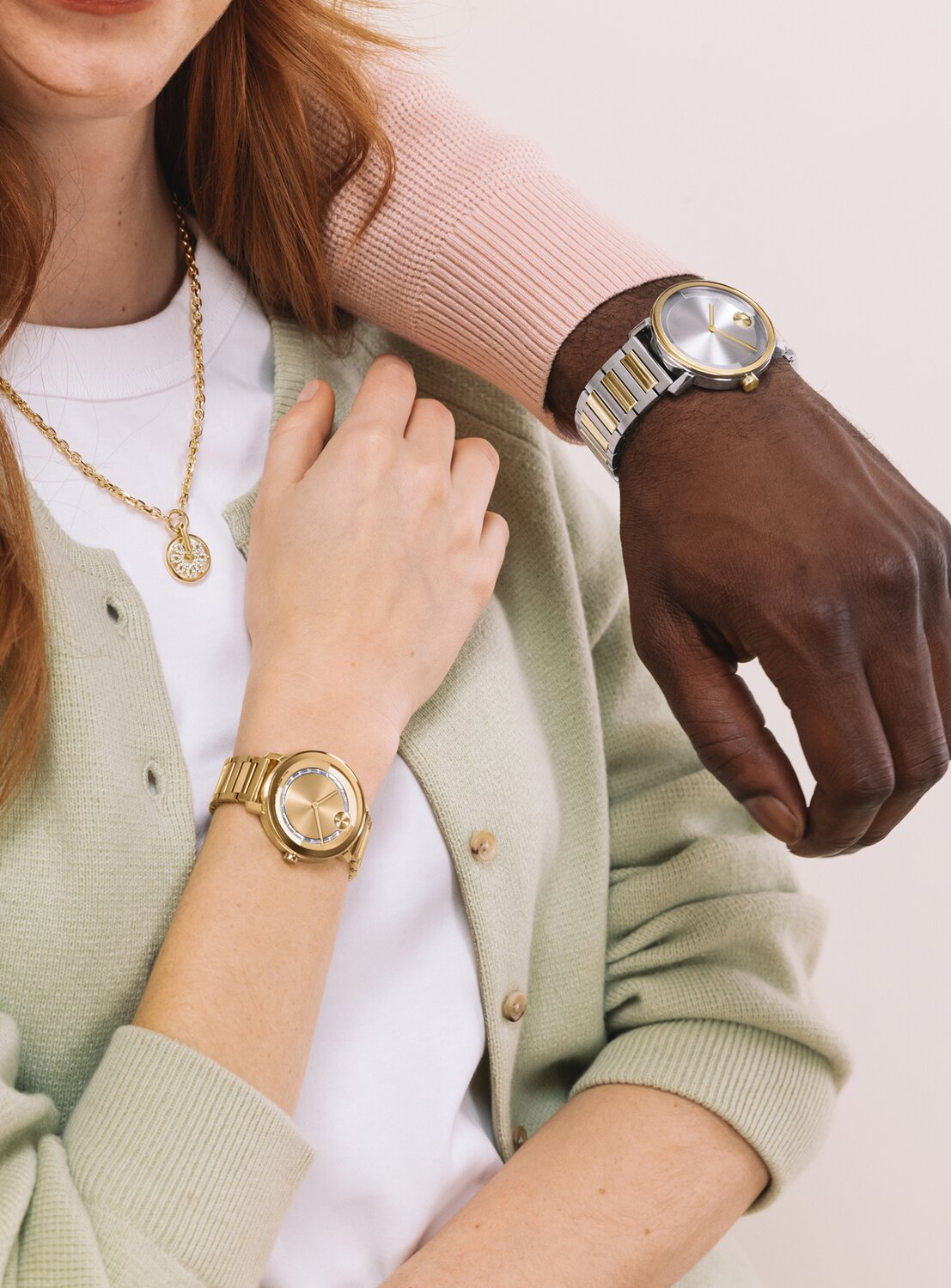 Man in a pink sweater resting his arm on a woman's shoulder while wearing a two tone BOLD Evolution 2.0 watch. The woman is wearing a green cardigan, the gold tone fleur necklace with lab grab down diamonds, and a gold tone BOLD Evolution 2.0 watch with baguette crystals on the dial. 