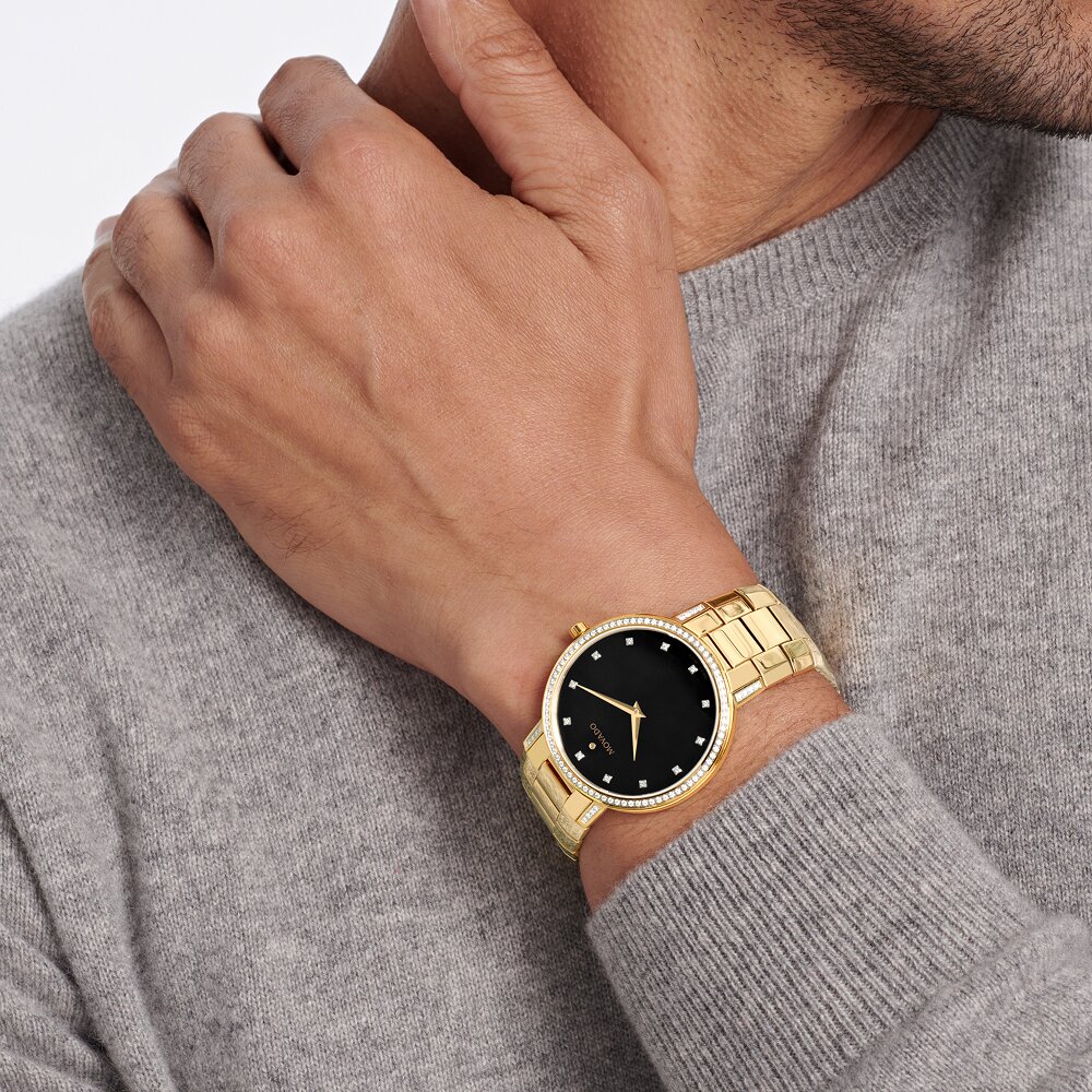 Man in a grey sweater with his hand on his shoulder wearing the Faceto watch with gold tone bracelet and black dial with diamonds.