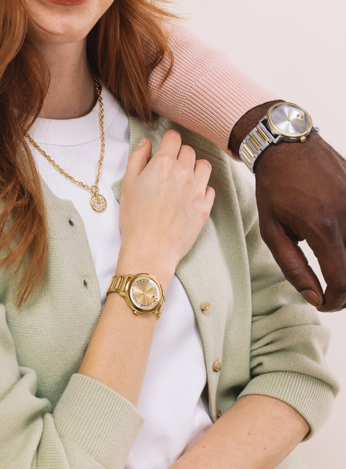 Woman in a green cardigan and white top wearing a gold tone BOLD Evolution 2.0 watch with baguette crystals and a gold tone fleur necklace with lab grown diamonds. A man in a pink sweater wearing the two tone BOLD Evolution 2.0 bracelet watch has his arm on her shoulder.