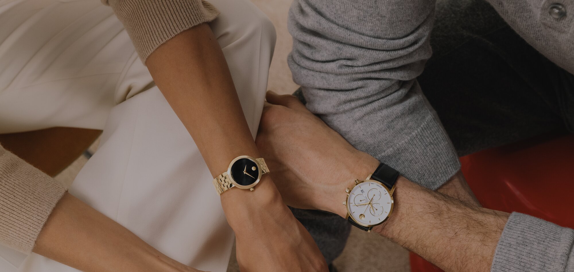 Woman wearing a gold tone Museum Classic bracelet watch with a black dial sitting next to a man wearing a Museum Classic Chronograph strap watch with white chronograph dial.