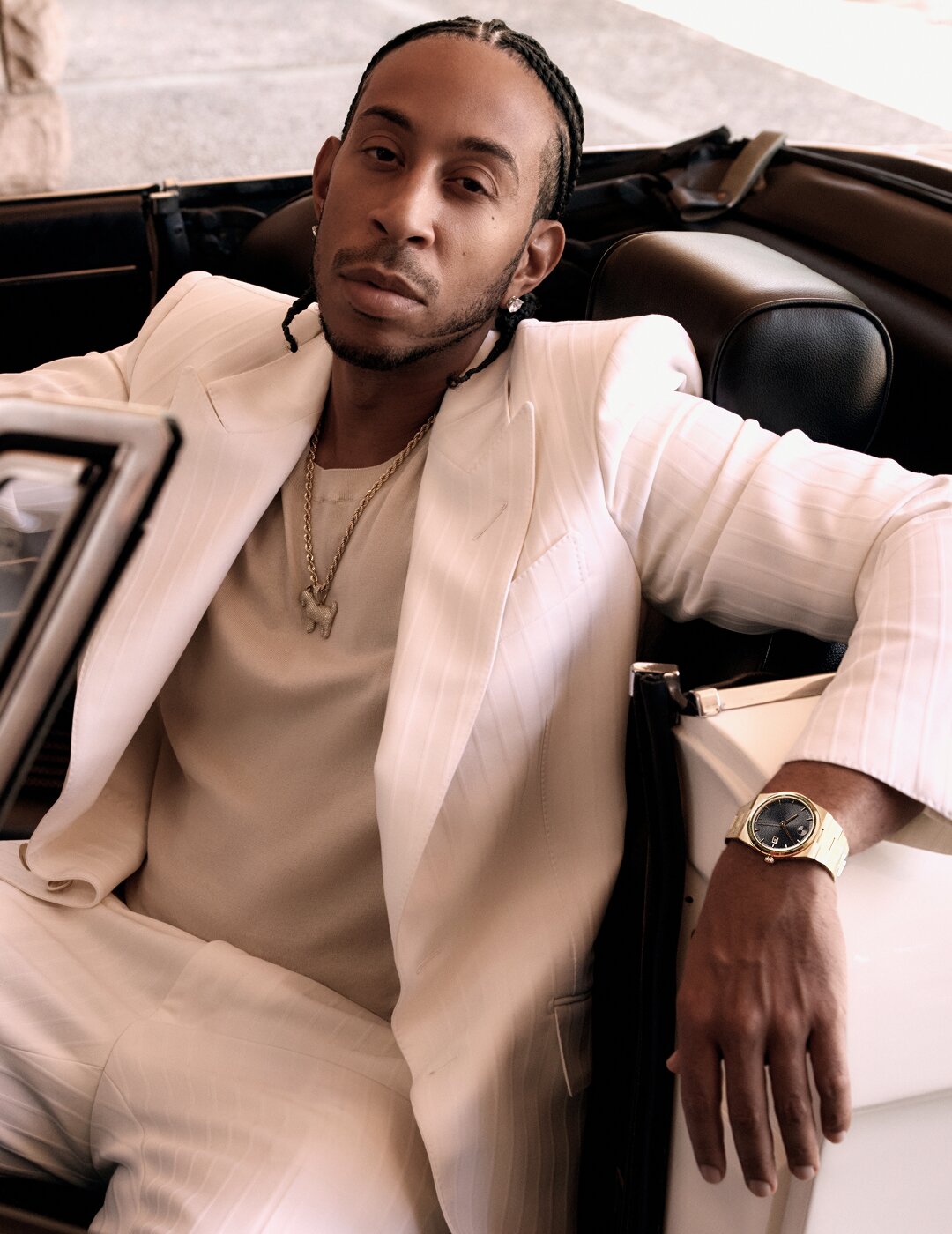 Ludacris sitting in a car, wearing a white suit and the BOLD Quest watch with gold tone bracelet and black dial.