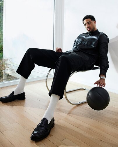 Tyrese Haliburton sitting in a chair, bouncing a black basket ball, wearing a black leather jacket, black pants, high white socks, black loafers and the Heritage 1917 watch with stainless steel bracelet and grey dial.