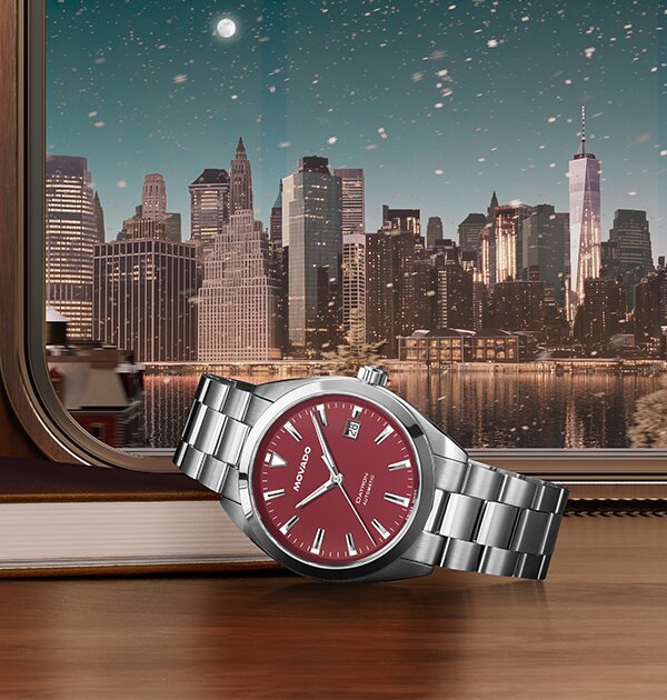 Silver watch with red dial laying on a book on a train with a cityscape in the background