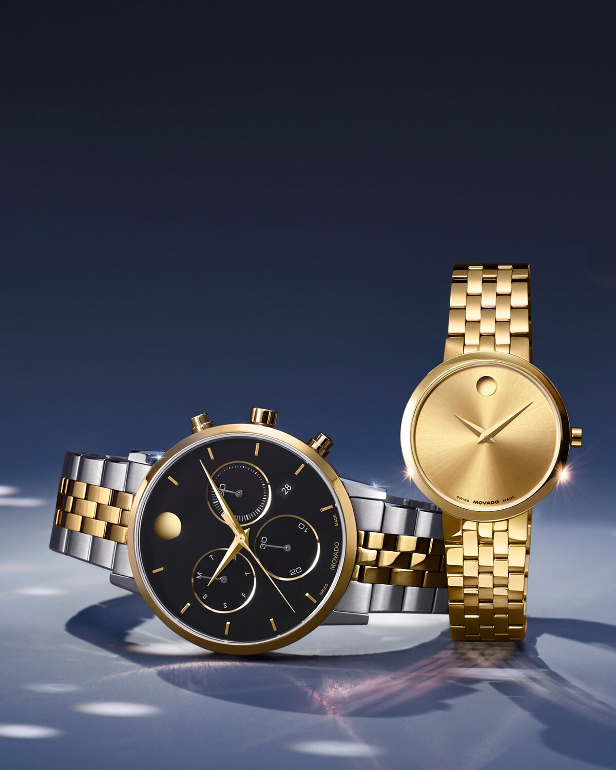 Two tone Museum Classic bracelet watch with black chronograph dial laying on its side. The women's gold tone Museum Classic bracelet watch is next to it.