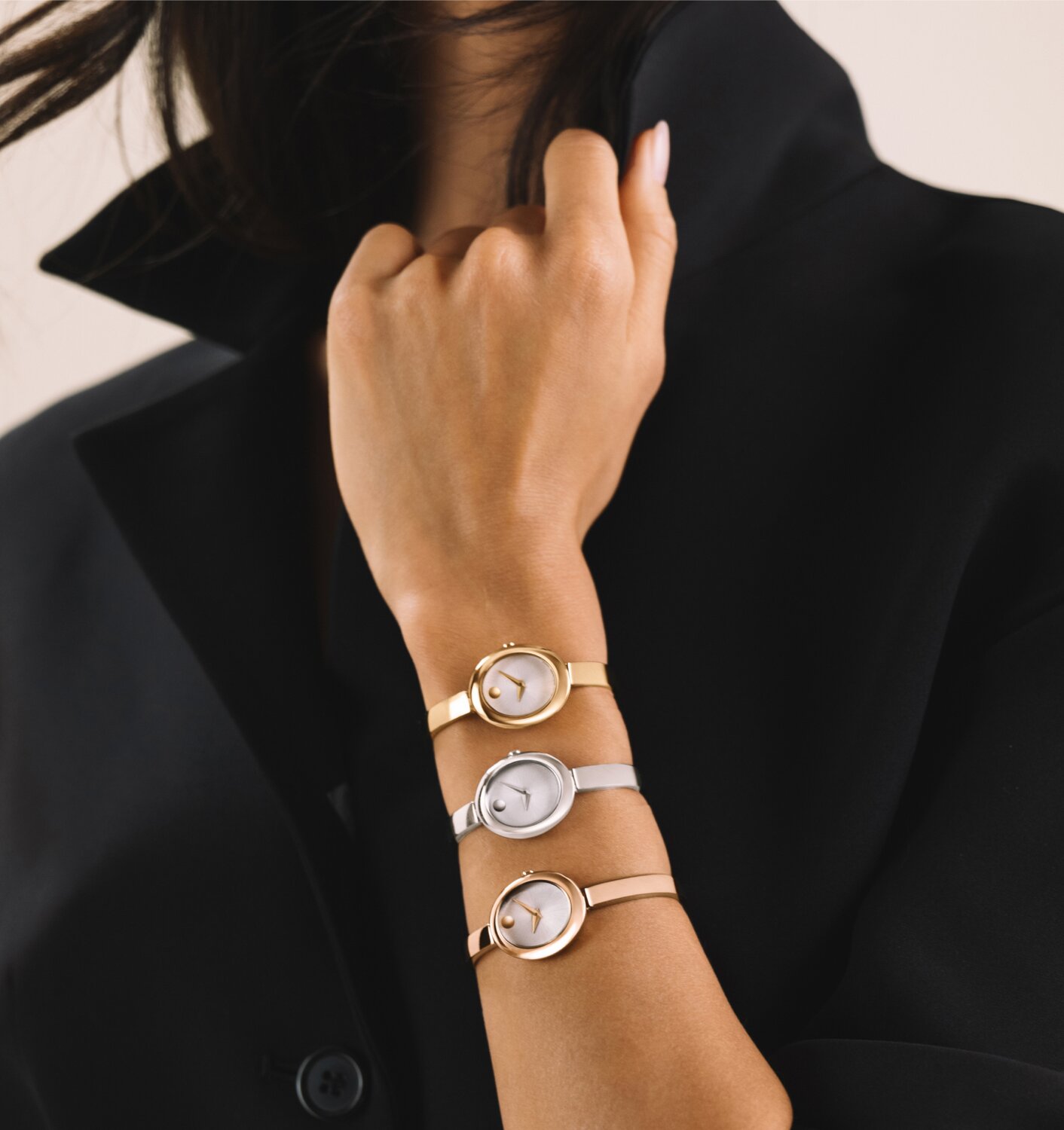 Woman in a black coat wearing a stack of three Oval Museum Bangles on her arm. Leading with gold tone, followed by silver, and ending with rose gold tone.