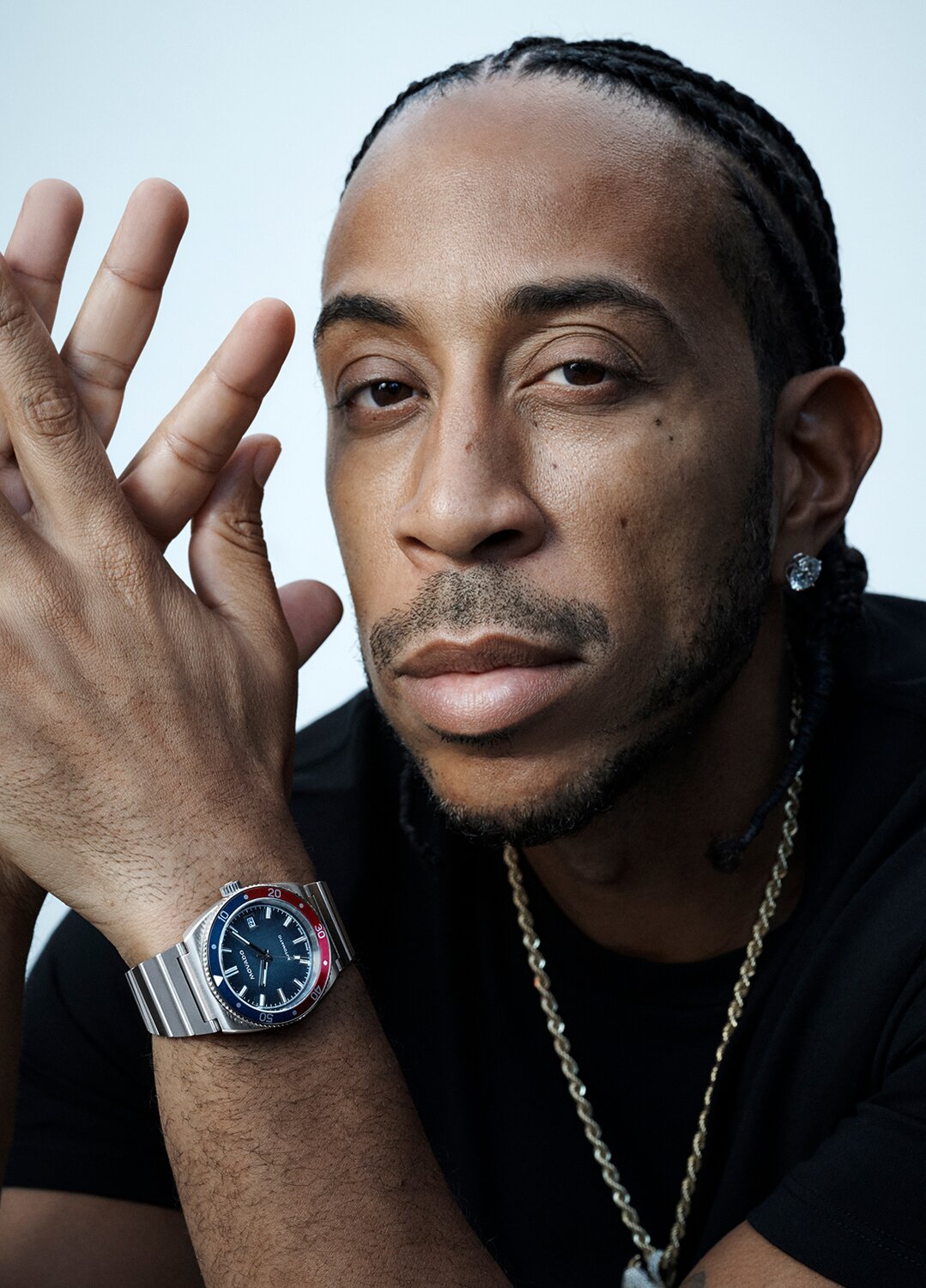 Ludacris in a black t-shirt with his hands crossed wearing an Ondoplan Automatic watch with silver bracelet and blue dial.