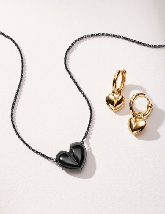 Black plated heart necklace next to gold plated hoop earrings with heart charm.
