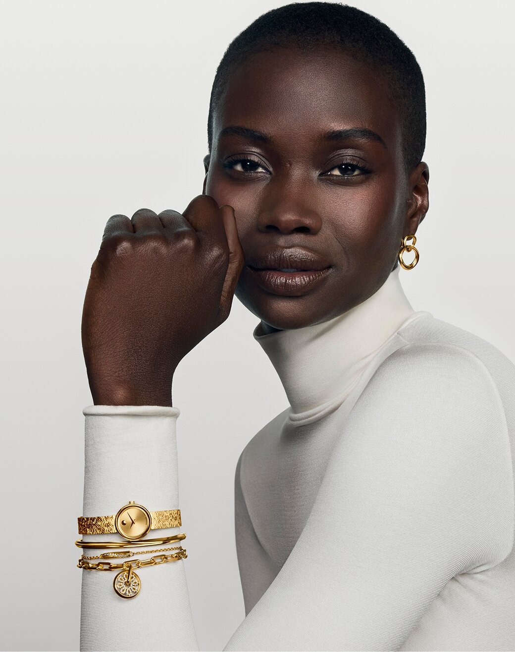 Woman in a white turtle neck wearing gold tone Movado earrings, bracelets and Museum Velura Min watch.