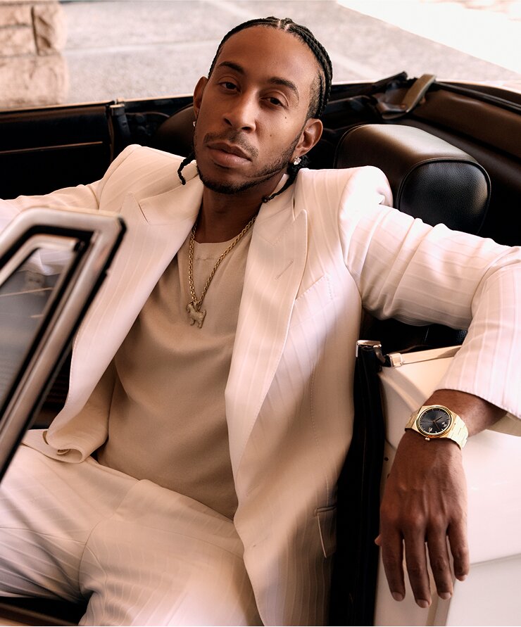 Ludacris wearing a white suit, sitting in a car, wearing the BOLD Quest watch with gold tone bracelet and black dial.