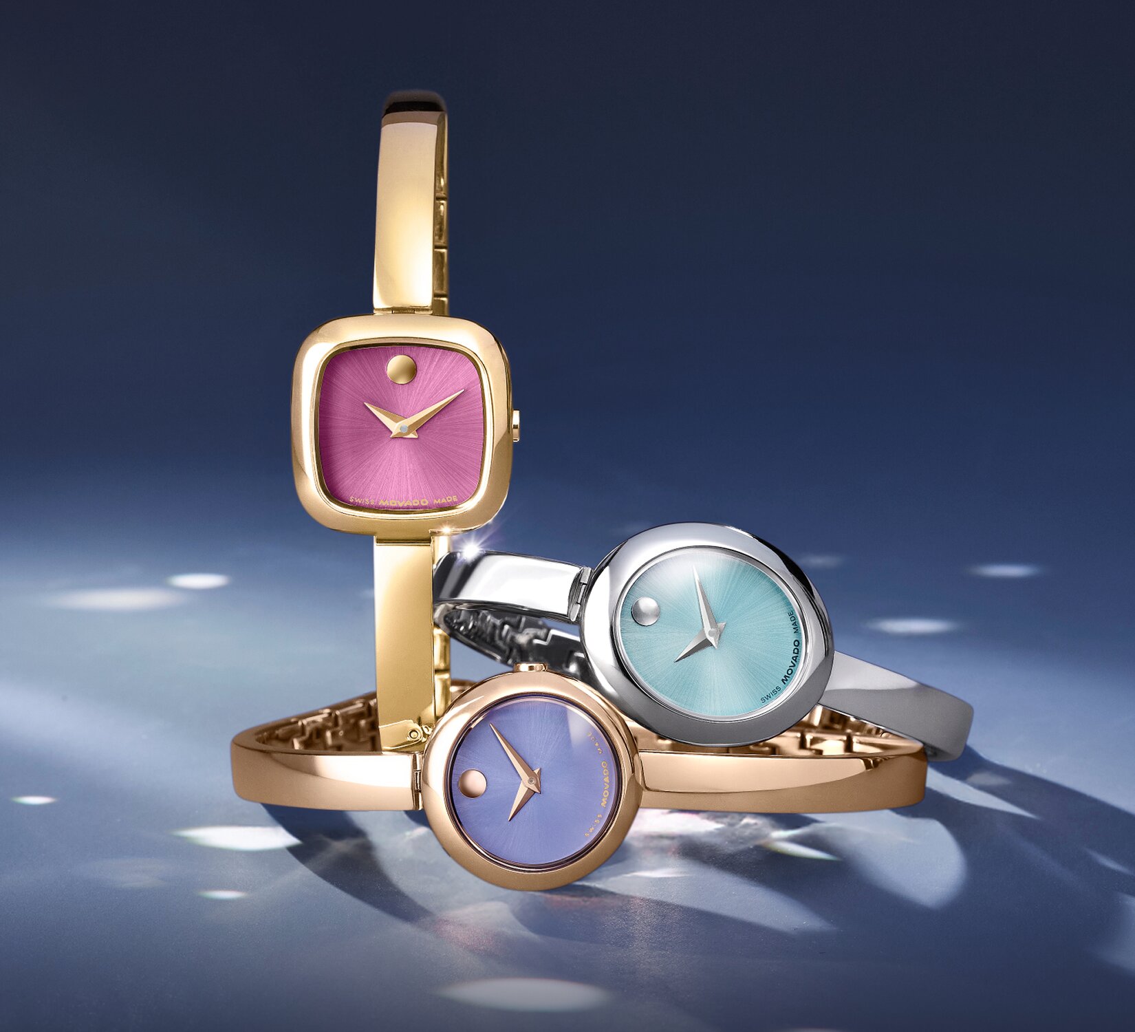 Museum Bangle watch with gold tone bracelet and pink square dial next to two Museum Bangle watches stacked on top of one another. The stainless steel Museum Bangle watch with blue oval dial is laying on top of the rose gold watch with purple dial.