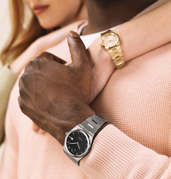 Man wearing a silver BOLD Quest Automatic bracelet watch with black dial holding a woman's hand that is wearing a gold tone BOLD Quest mini bracelet watch on her wrist