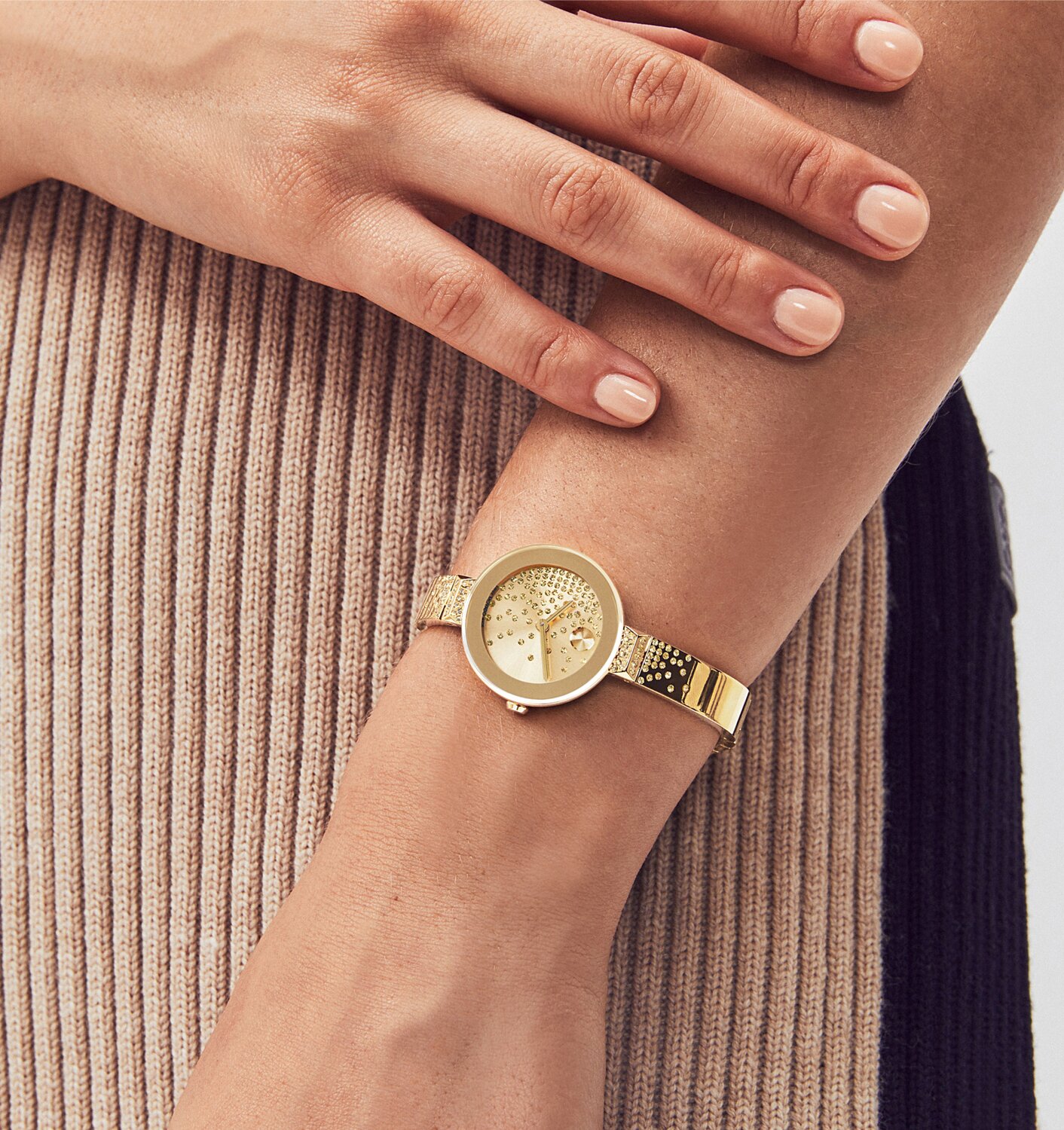 Woman in brown and black sweater dress wearing a gold tone BOLD bangle watch with crystal diamonds on the dial and bangle bracelet