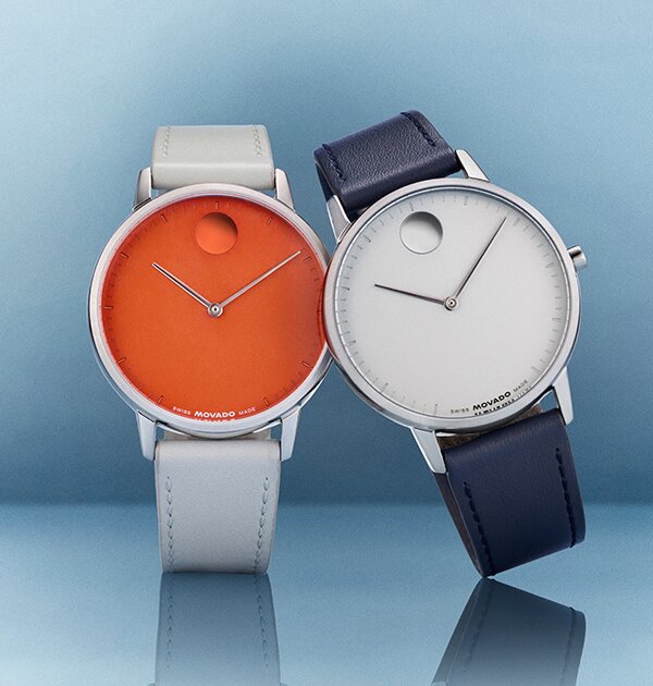 Silver strap Movado Face watch with orange dial next to a blue leather strap Movado Face watch