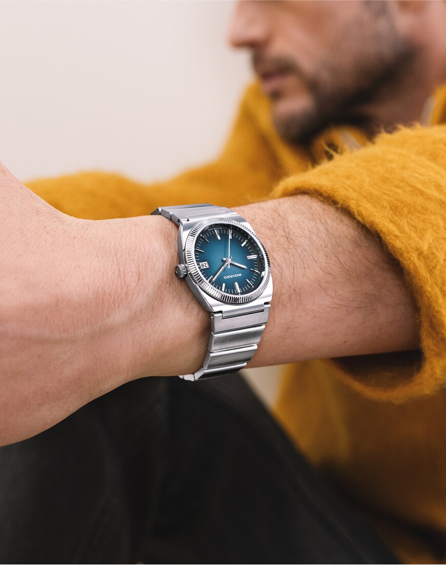 Museum Classic Automatic watch with stainless steel bracelet and black dial in front of a yellow background.