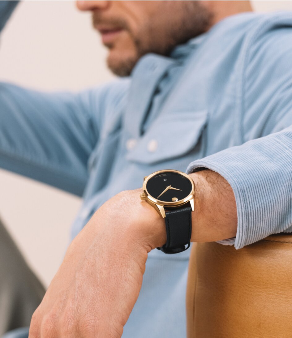 Man in a blue corduroy button down shirt wearing a Museum Classic Automatic watch with black leather strap and black dial with gold tone accents.