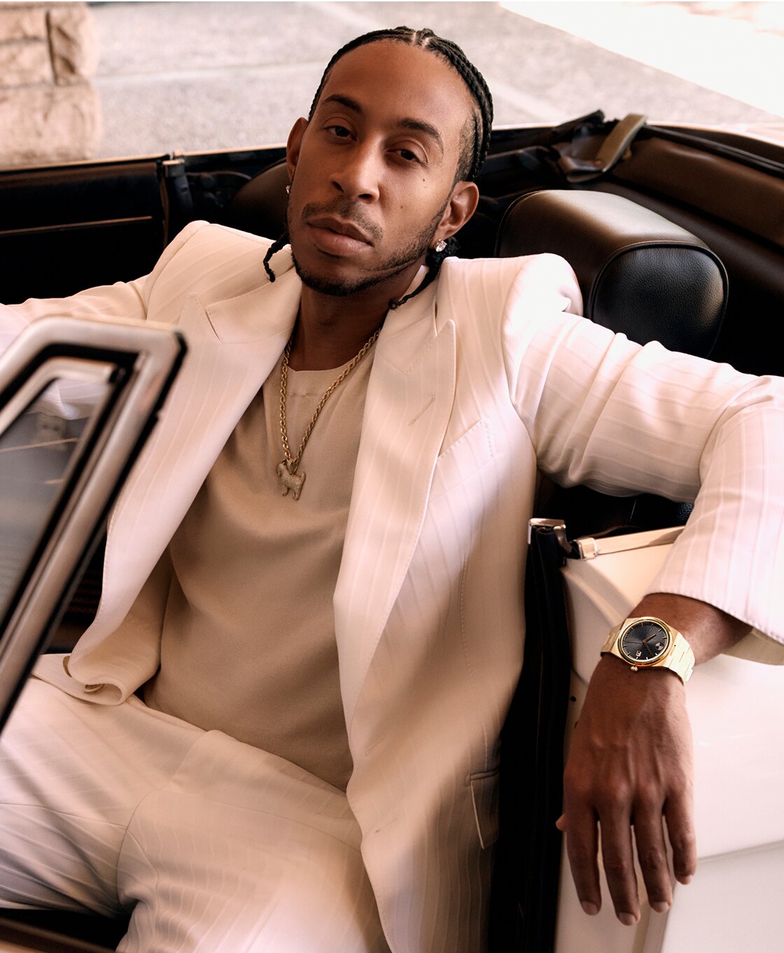 Ludacris wearing a white suit, sitting in a car, wearing the BOLD Quest watch with gold tone bracelet and black dial.