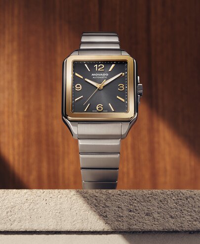 Heritage 1917 watch with stainless steel bracelet and grey dial sitting on stone in front of a wooden wall.