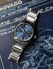Heritage 1917 watch with stainless steel bracelet and grey dial sitting on stone in front of a wooden wall.