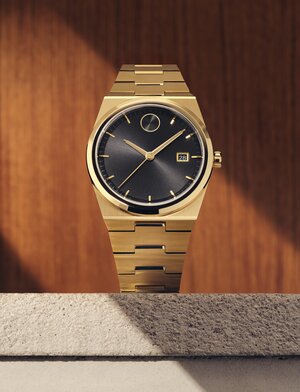 BOLD Quest watch with gold tone bracelet and black dial sitting on stone in front of a wooden panel.