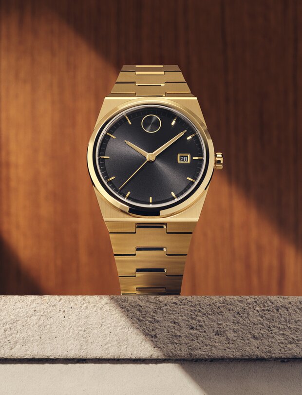 BOLD Quest watch with gold tone bracelet and black dial sitting on stone in front of a wooden panel.
